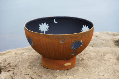 Tropical Moon 36-inch steel fire pit with palm tree and crescent moon cutouts, unlit, on a lakeside rock ledge.