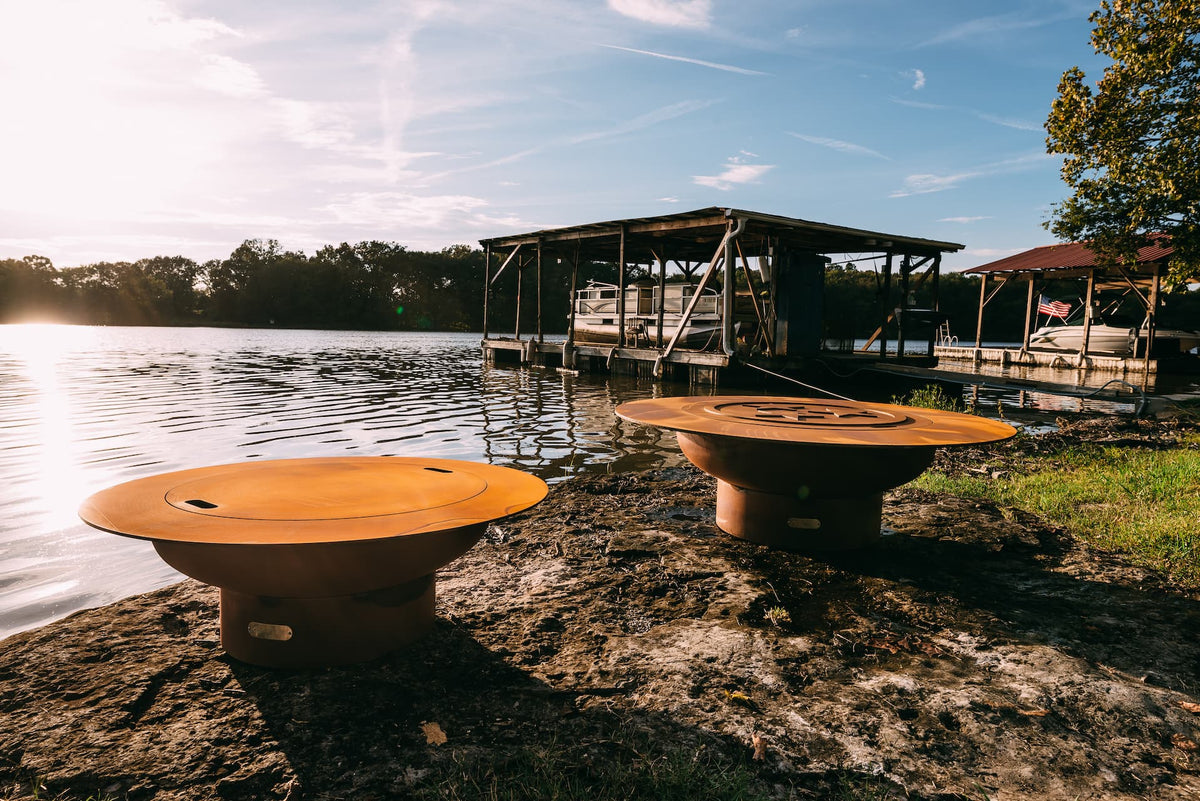 Two 54" Magnum carbon steel fire pits with lids on a lakeside with a dock and trees in the background.  Shows both sides of the lid.