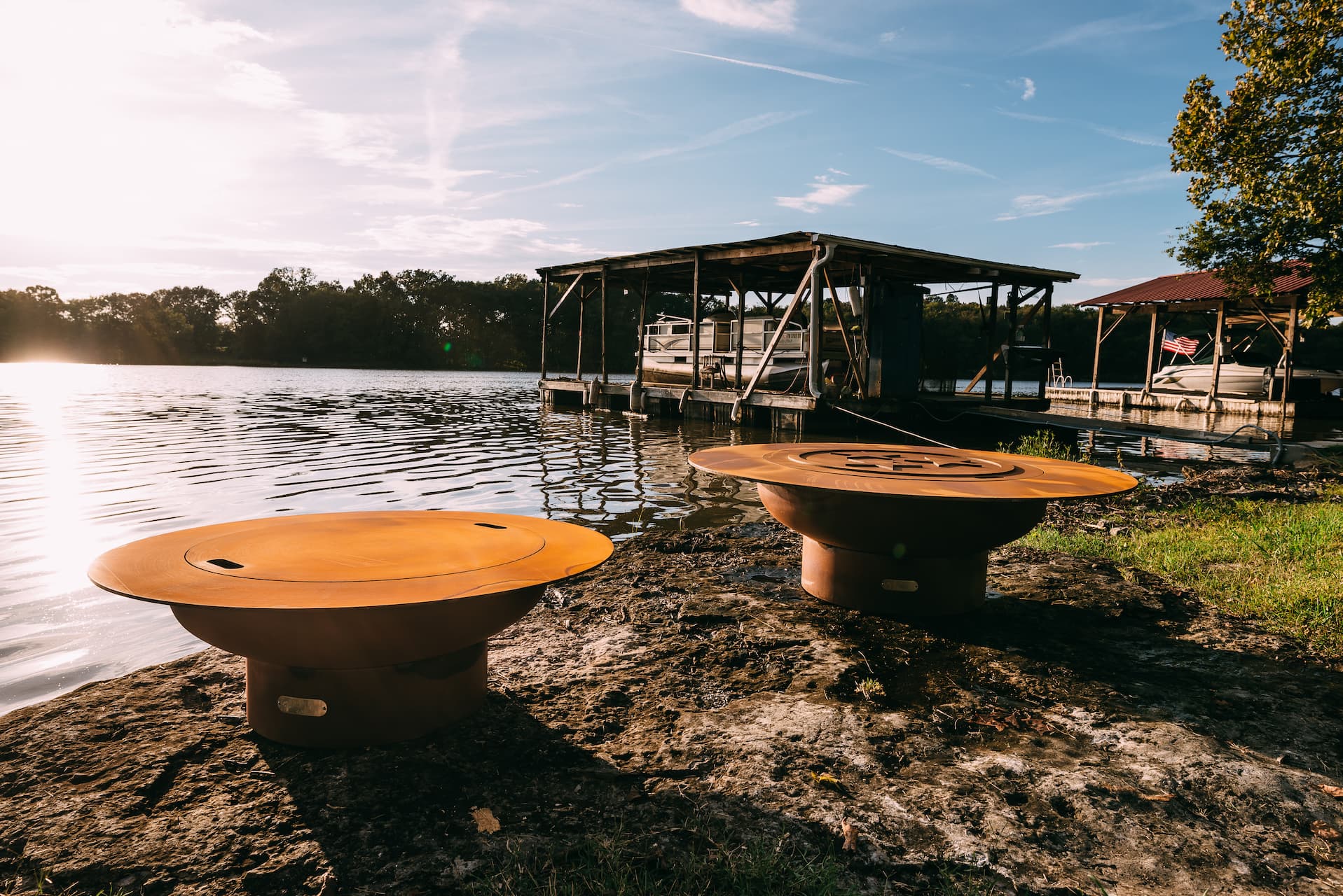 Two 54" Magnum carbon steel fire pits with lids on a lakeside with a dock and trees in the background.  Shows both sides of the lid.