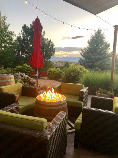 Wine barrel fire pit burning on an outdoor patio at sunset with wicker seating and string lights.